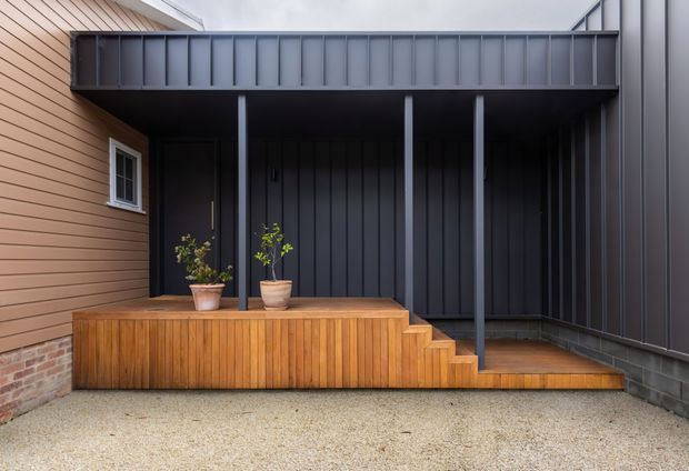 Contemporary residential courtyard or entry area with a raised timber deck and integrated timber-clad bench or planter. Dark charcoal metal cladding forms the covered porch and side wall, supported by slender steel posts, while a brick base and small white-framed window add contrast. Timber steps lead down to a pale gravel-paved surface, with potted plants providing subtle greenery.