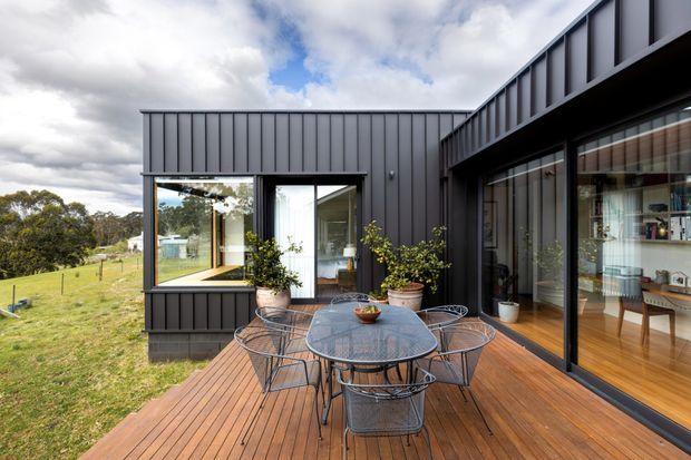 Exterior view of a contemporary single-storey home with dark vertical metal cladding, extensive glazing and a timber deck. The deck is furnished as an outdoor dining area with a round table and wire chairs, overlooking a grassy rural landscape. Interior dining and living areas are visible through the glazed doors.