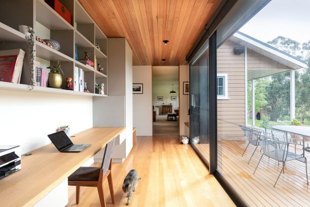 A bright, narrow open-plan interior combining a compact home office with a circulation and living area. Built-in white shelving and a timber desk line the left wall, while full-height black-framed glazing opens to a timber deck with outdoor dining furniture. The space features a warm timber-lined ceiling and matching timber floor, with a view toward a furnished living room beyond.
