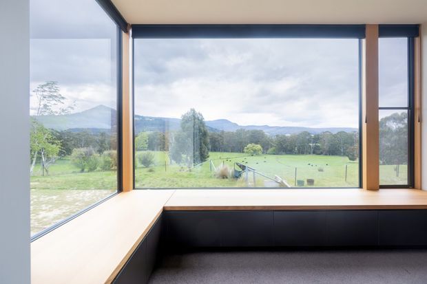 A quiet interior window seat or built-in viewing nook framed by expansive floor-to-ceiling glazing. Pale timber surrounds and bench surfaces contrast with dark fixed cabinetry below, while the windows overlook a rural green paddock, trees, and distant rolling hills. The space is simple, contemporary, and strongly connected to the landscape.