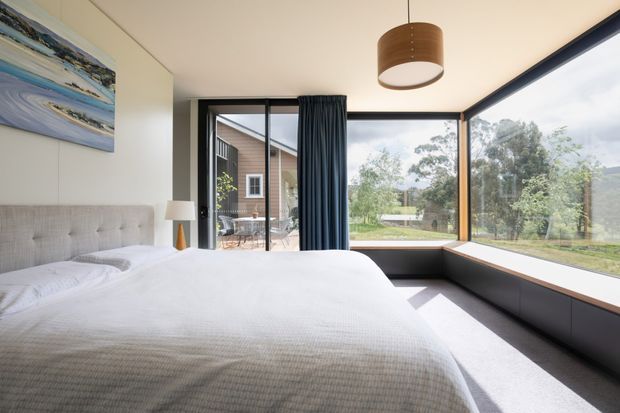 Bright contemporary bedroom with a large upholstered bed, neutral bedding, dark built-in window seating and storage, floor-to-ceiling corner glazing, dark curtains, and a simple timber pendant light. The glazing overlooks a leafy suburban landscape and an adjacent building, while a small outdoor terrace is visible beyond the door.