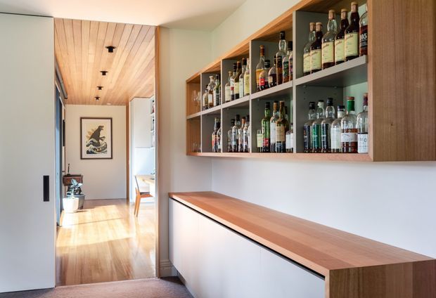 A warm, contemporary home bar occupies a built-in wall of open timber shelving stocked with bottles and glassware above a long timber-topped cabinet. The bar connects to a bright hallway or dining area, with pale walls, timber flooring, a timber-lined ceiling, and integrated ceiling spotlights. A framed artwork and potted plant are visible in the adjoining space.