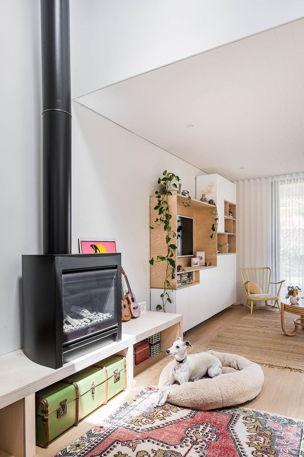 Bright contemporary living room featuring a black freestanding wood-burning stove with a prominent flue, custom white and timber built-in media and display cabinetry, pale timber flooring, a woven rug, and a seating area near sheer curtains. A dog bed and dog are visible in the foreground, with guitars, plants, and a yellow timber armchair adding character.