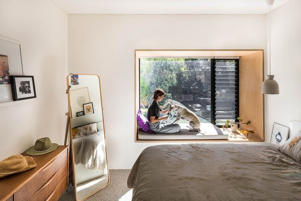 A bright, warmly styled bedroom with a large built-in window seat overlooking greenery. The room features a low bed with taupe bedding, light neutral walls, grey carpet, timber bedside/storage furniture, a full-length leaning mirror, framed artwork, and a pendant light. A person and dog sit on the window seat, while black louvres provide privacy and sun control.