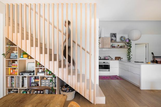 Bright open-plan residential interior showing a timber staircase with vertical timber balustrade and integrated under-stair shelving, opening into a contemporary white kitchen. A timber dining table occupies the foreground, with light timber flooring, white cabinetry, open shelving and indoor greenery.