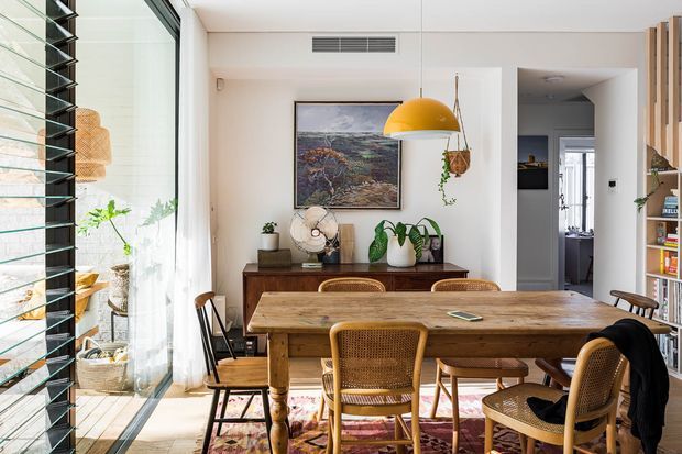 Bright, warmly styled dining room with a large timber dining table and woven cane chairs. The room features expansive floor-to-ceiling glazing with horizontal louvres, white walls and ceiling, timber flooring, a colourful area rug, indoor plants, artwork, open shelving, and a mustard-yellow pendant light. A hallway and adjacent spaces are visible beyond the dining area.