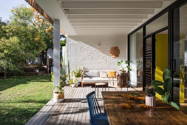 Sunlit covered outdoor deck and patio adjoining a contemporary home, with timber decking, a large timber dining table, black chair, outdoor sofa, potted plants, and a white-painted brick feature wall. Black-framed glazing opens toward the interior, where a vivid yellow wall is visible. The space overlooks a leafy garden and has exposed painted ceiling beams, creating a relaxed indoor-outdoor entertaining area.