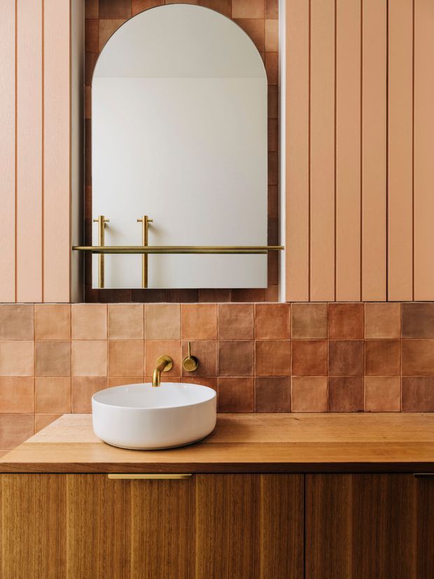 A warm, carefully detailed bathroom vanity with a white vessel basin set on a timber benchtop and dark timber cabinetry. The wall combines square terracotta-toned tiles with vertical peach-toned panelling. A large arched mirror is framed by darker tiles, with brass wall-mounted tapware and a brass horizontal rail adding refined accents.