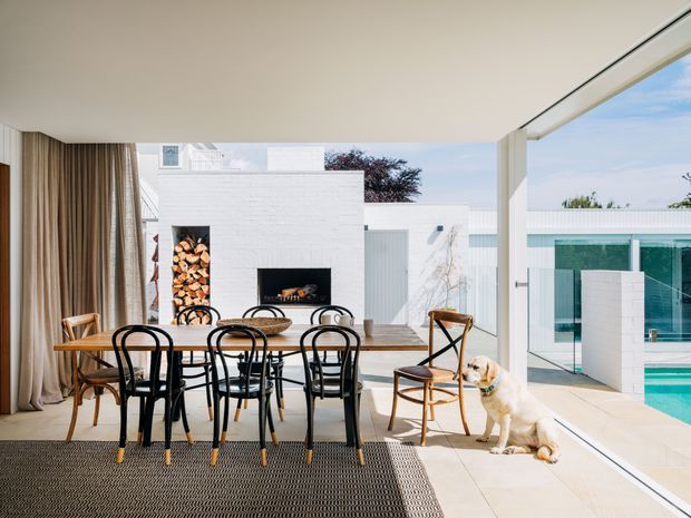 Bright open-plan dining area opening directly onto a pool terrace through extensive floor-to-ceiling glazing. The room features a long timber dining table with black bentwood-style chairs, a woven rug, full-height neutral curtains, and a white masonry fireplace with integrated firewood storage. A modern white poolside structure is visible outside, with a dog near the glazing.