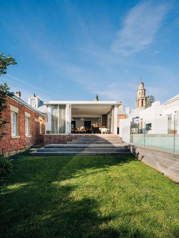 Exterior view across a grass courtyard toward a contemporary rear extension with broad steps, an open-plan glazed living area, pale rendered walls, brick boundary walls, and a raised pool or terrace edge with glass balustrading. A historic tower is visible beyond the neighbouring buildings, creating a contrast between the modern addition and the established urban context.