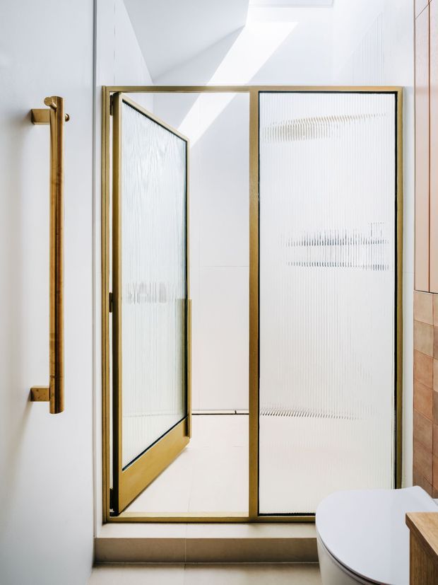Bright contemporary bathroom featuring a gold-toned framed frosted or reeded-glass shower enclosure with a hinged door. White walls and floor create a clean, airy setting, with a warm timber-toned tiled wall and partial vanity visible at right. A large vertical brass/gold door handle is prominent on the left.