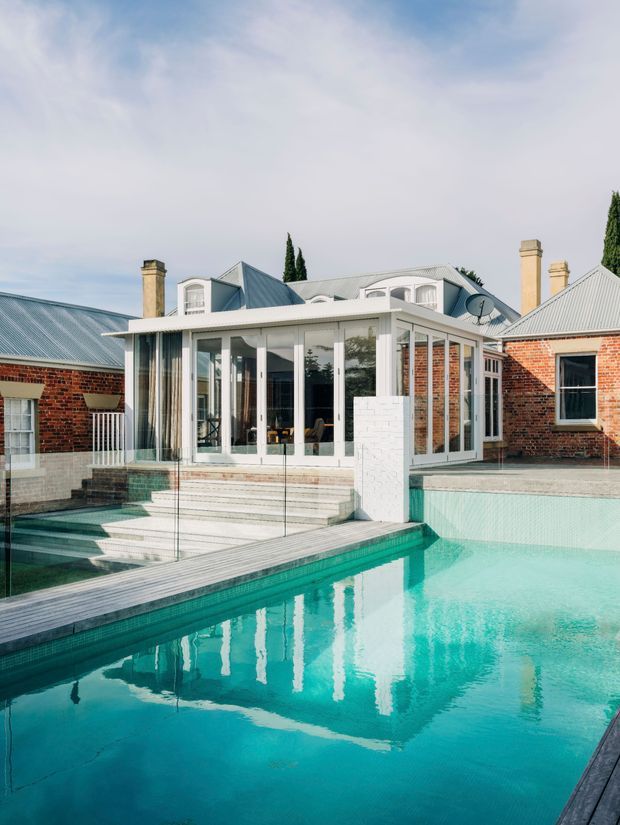 Exterior view of a traditional brick residence with a glazed, white-framed pavilion or sunroom opening directly onto a turquoise swimming pool. The house features corrugated metal pitched roofs, multiple brick chimneys, dormer windows, and a glass pool barrier. Pale timber decking and stepped access lead into the pool, with the glazed extension forming the visual focus.