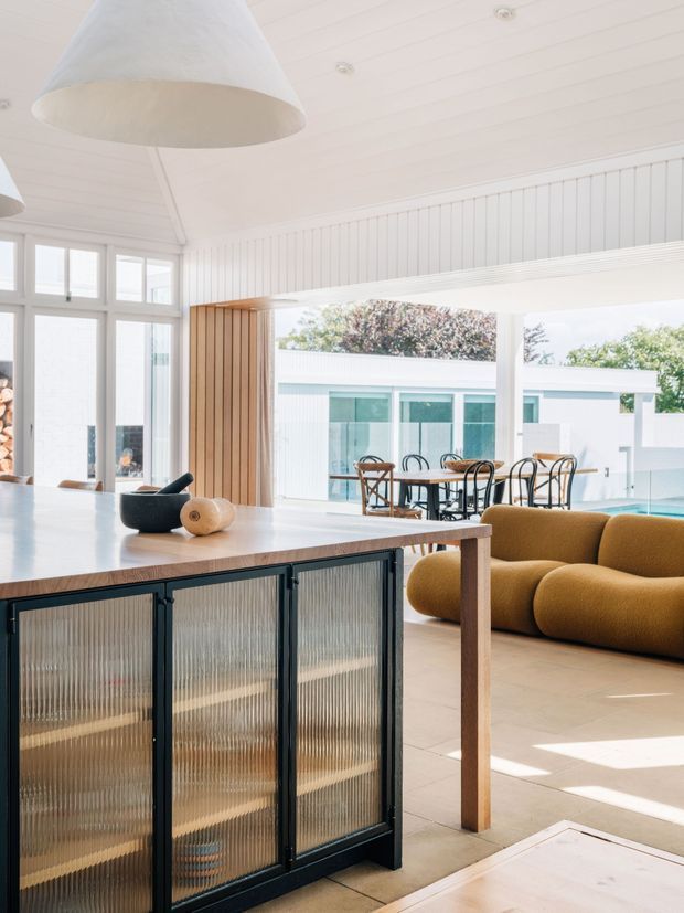 Bright, open-plan contemporary interior centred on a large kitchen island with a timber benchtop and black-framed ribbed translucent-glass cabinetry. The space has white vertical wall panelling, a white lined ceiling, expansive glazing, timber accents, sculptural pendant lights, and a mustard upholstered sofa. A dining area and an outdoor pool courtyard are visible beyond the open living space.