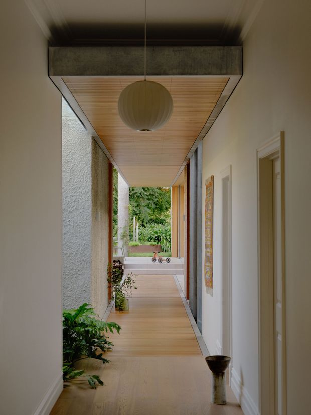 A narrow, refined residential hallway leading toward a planted outdoor courtyard. The corridor features warm timber flooring, a timber slatted ceiling soffit, white textured walls, dark structural or metal framing, and a large suspended spherical pendant light. Openings along the passage reveal greenery and a small outdoor water feature or pool beyond, while artwork and potted plants add visual interest.