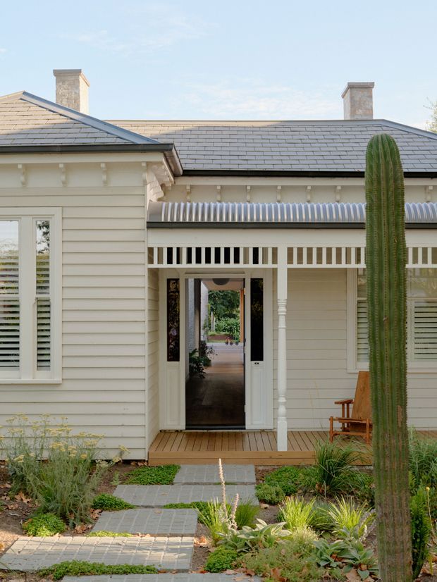 Front exterior view of a single-storey light-painted weatherboard house with a low-pitched tiled roof, two chimneys, decorative eaves brackets, a covered entry porch with fretwork ventilation panels, and an open central doorway. A paved garden path leads through drought-tolerant landscaping with a tall columnar cactus and ornamental grasses.