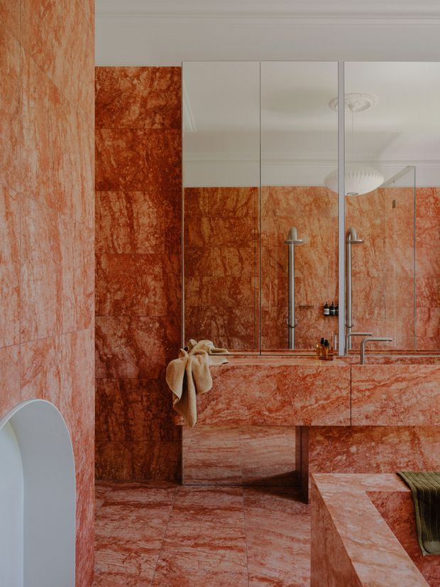 A compact, richly finished bathroom clad extensively in veined red-orange stone or marble-look slabs across the walls, floor, vanity, and bath surround. A large mirrored cabinet spans the vanity wall, with a wall-mounted basin tap, shower hardware, and a freestanding or partially visible white toilet at left. The space has a sculptural, highly cohesive monochromatic material palette with polished metal fixtures and soft ceiling lighting.