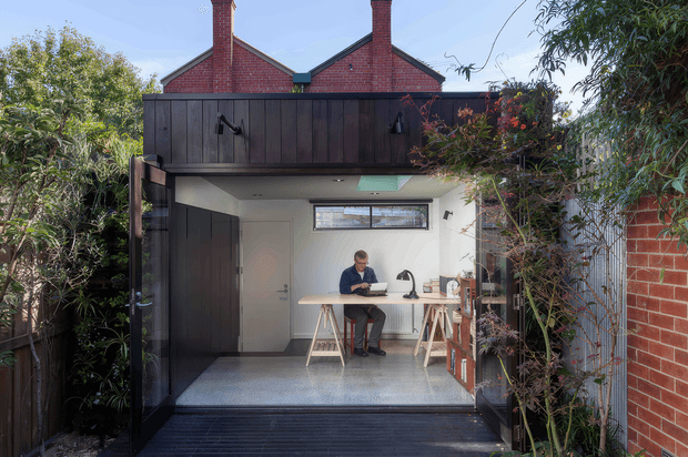 A compact, contemporary backyard studio or workspace viewed from an outdoor deck through wide-open dark timber doors. Inside, a person sits at a simple timber desk with a laptop, task lamp and small furnishings. The studio has white walls, a polished grey concrete floor, a narrow horizontal clerestory window and a roof skylight. The exterior combines dark vertical timber cladding, exposed red brick walls and dense garden planting.