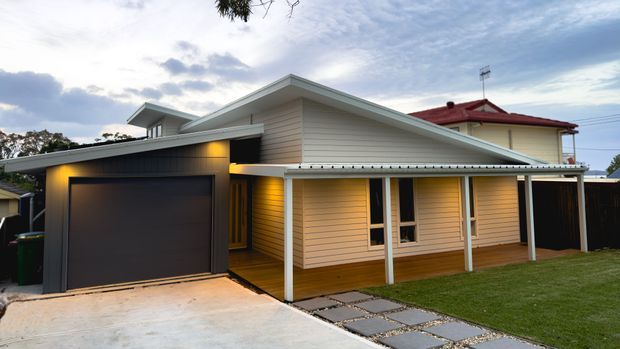 Exterior view of a single-storey suburban house featuring a contemporary gabled extension, pale horizontal weatherboard cladding, a dark attached garage, and a covered front verandah. A concrete driveway and stepping-stone path lead across a neatly landscaped lawn. The image also shows part of an older cream-coloured house with a red roof behind the extension.