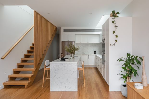 A bright, contemporary open-plan kitchen viewed from the adjoining living area. The space features a white island with a veined stone benchtop, white flat-panel cabinetry, a pale stone or marble-look splashback, warm timber flooring, and a timber open-riser staircase with vertical slatted screening. A skylight or roof window brings in natural light, while indoor plants and simple timber furniture add warmth.