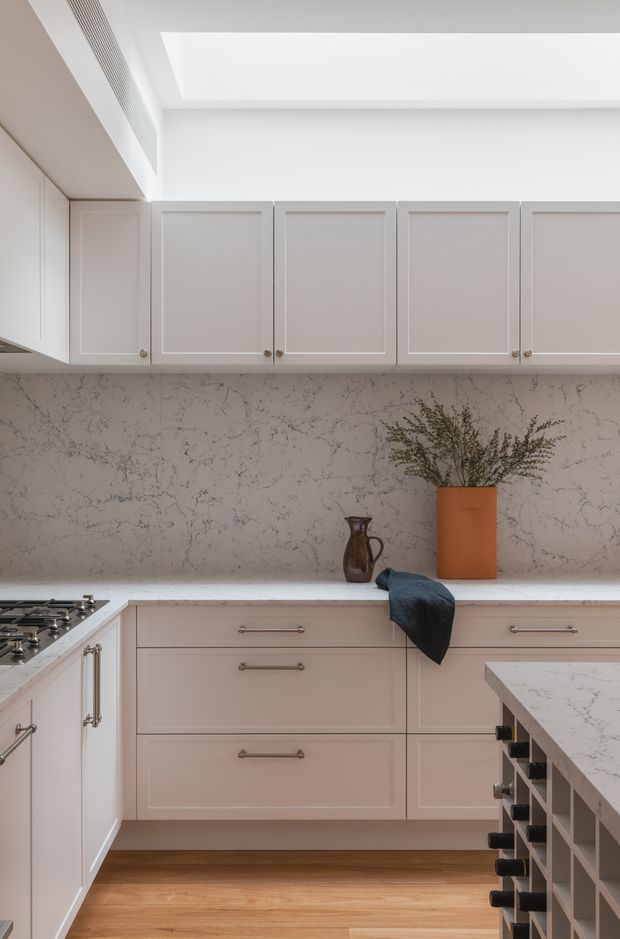 A refined, light-toned kitchen with pale framed cabinetry, a veined stone splashback and benchtop, integrated open shelving, and warm timber flooring. A gas cooktop is visible at left, while a partial island with bottle storage appears at right. The room is softly lit by a large ceiling skylight or recessed lightwell and features restrained decorative styling.