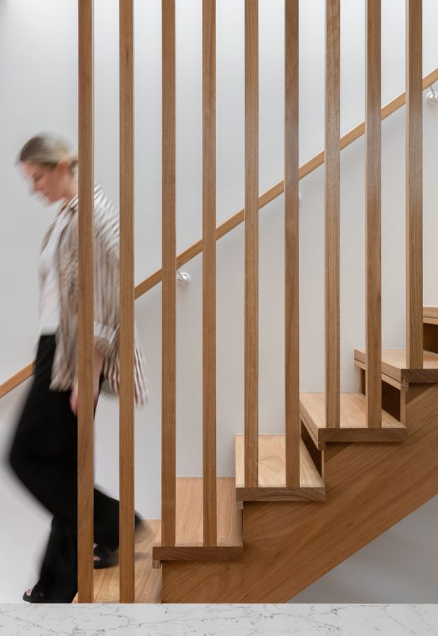 Interior view of a contemporary timber staircase with open risers and prominent vertical timber balusters forming a screen. The stair has warm wood treads, stringer and handrail against pale walls, with a blurred person passing beside it and a marble-look surface in the foreground.