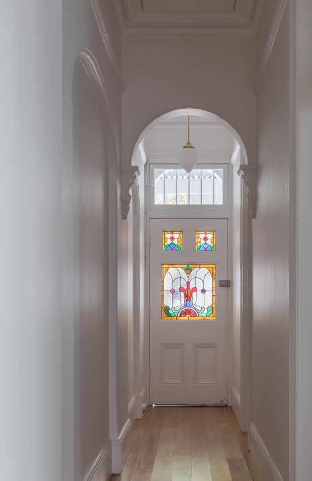 A narrow, elegant residential hallway leading to a white front door with distinctive multicoloured stained-glass panels. The corridor features white walls, decorative ceiling mouldings, ornate arched openings with classical trim and corbels, white timber skirting, and a warm timber floor. A simple pendant light hangs above the entry, creating a refined heritage character.
