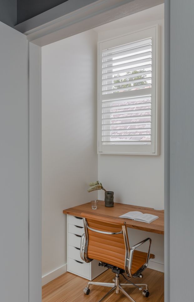 A compact home office nook with a warm timber desk, white drawer unit, and a vintage-inspired tan leather swivel chair. A white plantation-shutter window provides soft natural light, while simple decor and an open book create a quiet, refined workspace. White walls and trim contrast with the timber flooring and desk.