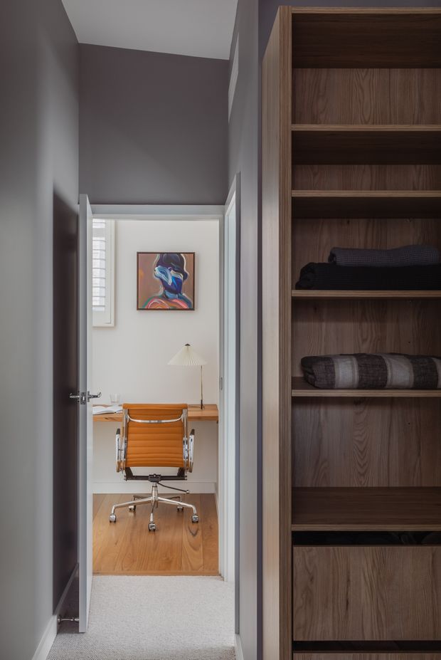 A narrow, softly lit hallway view toward a compact home office, with a built-in dark timber wardrobe or storage shelving dominating the right side. The office contains a timber desk, orange upholstered task chair, desk lamp, framed artwork, and warm timber flooring. The hallway has grey painted walls, white trim, and light carpet.