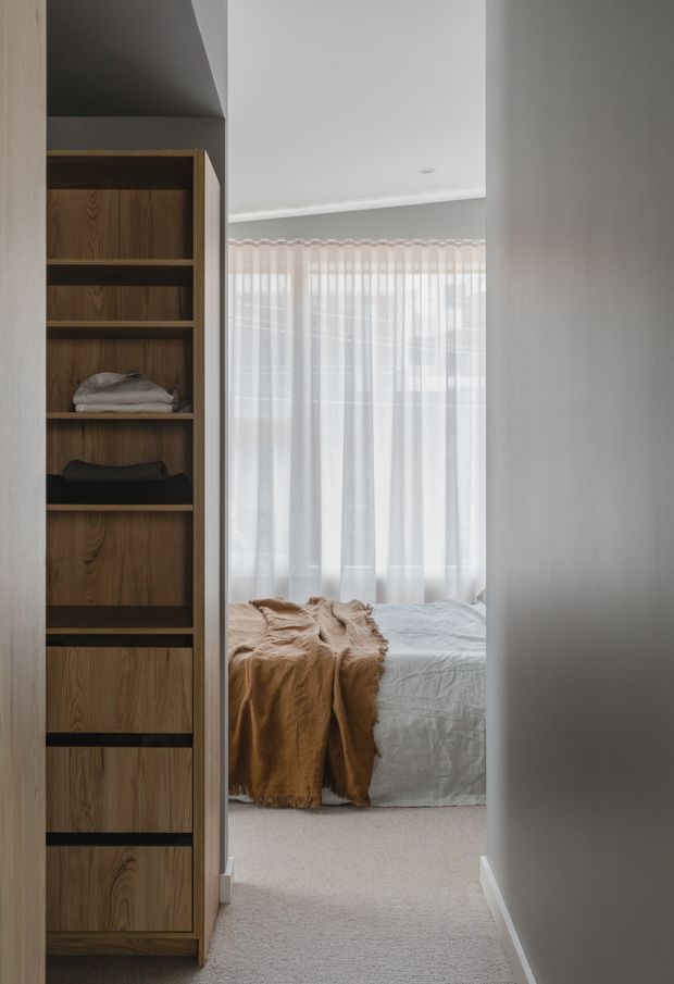 A view through a narrow hallway into a softly lit bedroom. The bedroom contains a neatly made bed with white bedding and a warm tan throw, sheer full-height curtains over a bright window, and neutral carpet. A dark mid-tone timber built-in shelving and wardrobe unit lines the left side, while simple white walls and ceiling create a calm, understated interior.