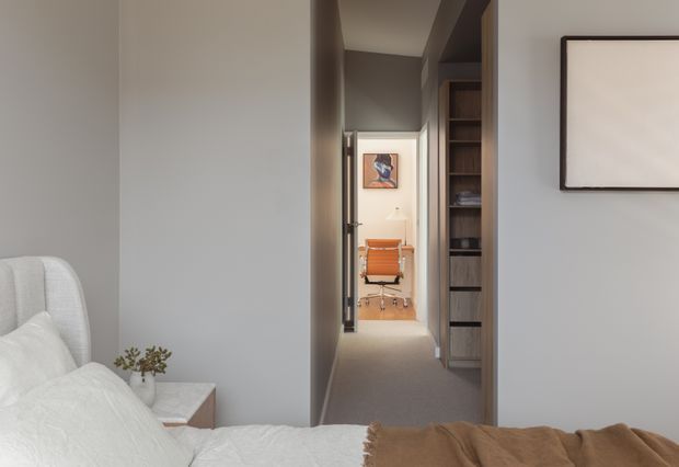 View from a softly styled bedroom toward a narrow hallway and compact home office. The foreground shows a light upholstered bed with white bedding and a brown throw, while the hallway includes built-in dark wood shelving and drawers, leading to a bright workspace with an orange office chair and wall art. The palette is neutral and warm, with clean contemporary detailing and subdued natural light.