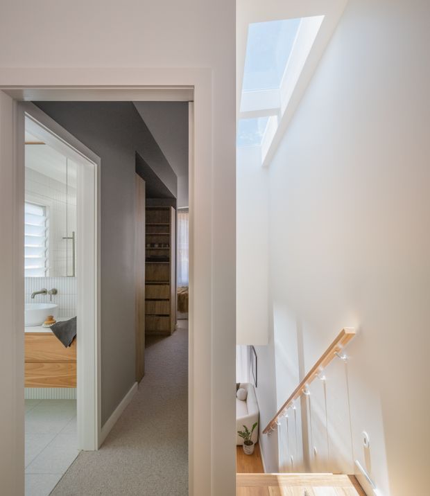 Bright interior view of a residential hallway and stair landing. The image looks along a carpeted corridor toward built-in timber shelving, with an open bathroom visible on the left containing a timber vanity and white basin. On the right, a timber staircase with a light wood handrail and clear glass balustrade descends beside a tall white wall with roof skylights. The space has a clean, contemporary, light-toned design.