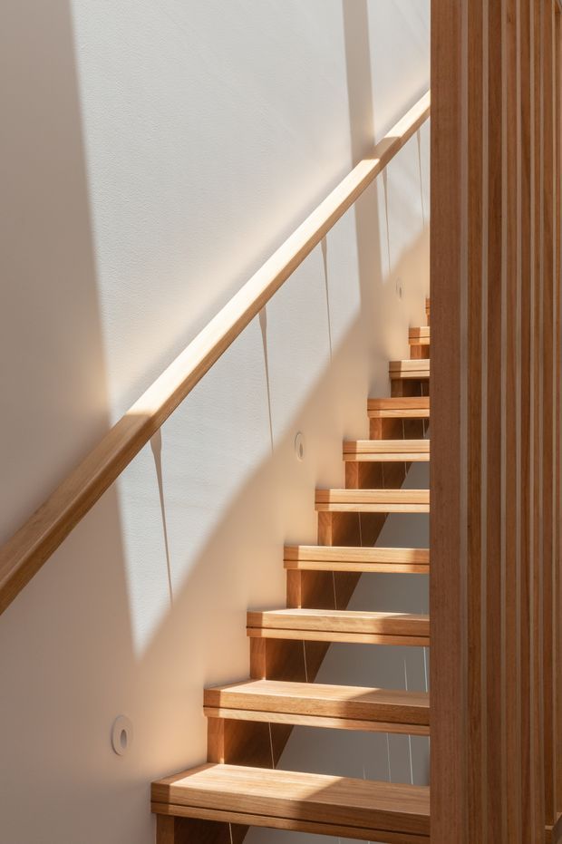 A narrow, sunlit interior stairway with open-riser timber steps, a light timber handrail, and thin cable balustrade. White plaster walls create strong diagonal shadows, while a vertical timber slat screen lines the right side. The design is simple, warm, and contemporary with natural daylight emphasizing the wood grain.