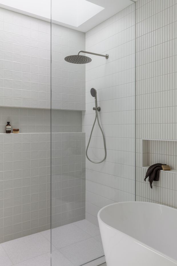 A bright, minimalist bathroom featuring a walk-in glass shower with large rain showerhead and handheld fixture, white tiled walls, recessed niches, a skylight, and a partially visible freestanding white bathtub. The space uses clean geometric lines and brushed metal fittings.