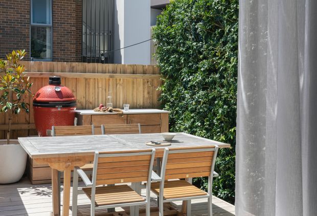 A compact, contemporary outdoor dining terrace viewed from beside a sheer curtain. The space features a timber dining table with matching slatted timber-and-metal chairs, a built-in timber outdoor cabinet or counter, and a red ceramic barbecue. Timber fencing, large leafy screening plants, concrete architectural elements, and a modern brick-and-metal building surround the terrace.