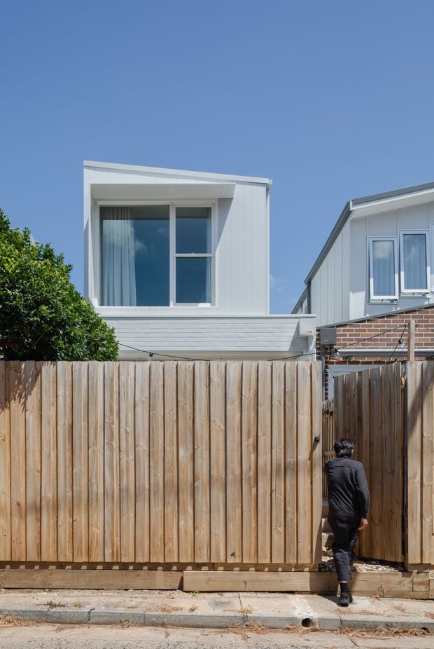 Exterior view of a contemporary multi-storey residential building behind a tall timber privacy fence. The main structure has white vertical cladding, a projecting upper-level box with large glazed windows, and a flat roofline. Part of an adjacent white-clad and red-brick dwelling is visible at right. A person is entering through a narrow timber gate, with paving and a small strip of street in the foreground under a clear blue sky.