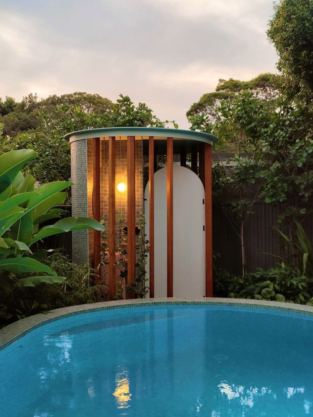 A compact curved poolside structure set within dense tropical landscaping. The rounded pavilion has a shallow curved roof with a green edge, vertical timber screens, translucent or glazed sections, small-format tiled wall surfaces, and a distinctive arched white door. A bright interior light glows through the screened facade, while the foreground is dominated by a blue swimming pool. The image has a lush, tranquil, softly lit atmosphere.