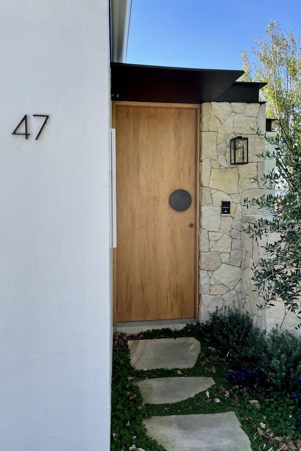 Exterior entry featuring a tall natural timber pivot door set into a pale irregular stone wall, beneath a dark projecting flat canopy. A white rendered wall displays the house number 47, with a black wall lantern, keypad, and landscaped stepping-stone path leading to the entrance.