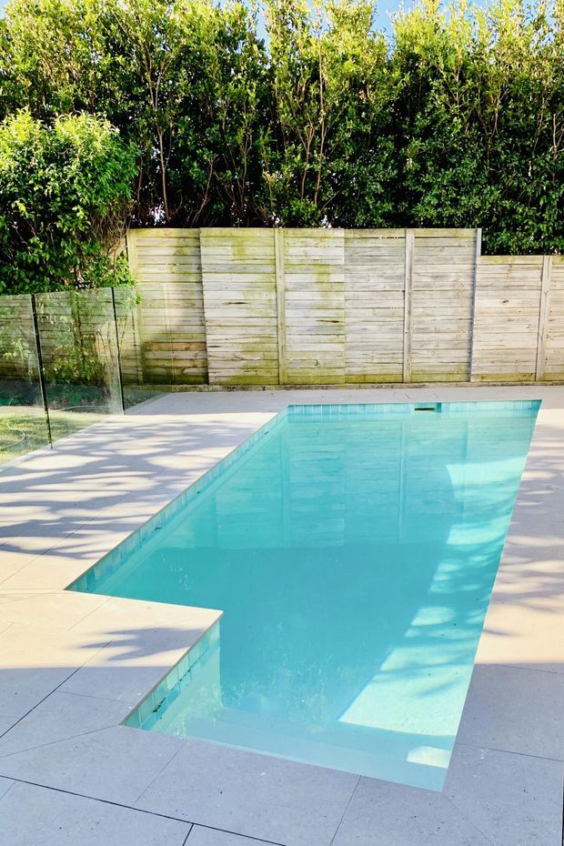 A bright outdoor swimming pool in a landscaped residential setting, surrounded by pale concrete or stone paving, a frameless glass safety fence, a weathered timber boundary fence, and dense green hedging. The rectangular pool has turquoise water and a shallow stepped entry area.