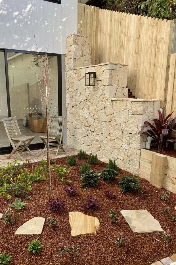 A landscaped courtyard garden beside a contemporary building, featuring a textured natural-stone privacy wall with integrated steps and a wall light, timber fencing and raised planter edging, mulched planting beds, and pale stone stepping slabs. A large black-framed glazed opening reveals an interior space, while timber folding chairs sit on a small paved area outside.