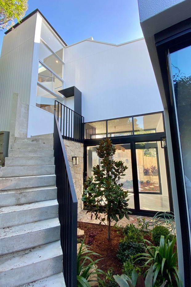 A bright contemporary courtyard viewed from within the house, featuring an exposed concrete staircase with black metal balustrade, white vertical-panel walls, natural stone cladding, and extensive black-framed glazing. A planted garden courtyard sits below the stair and beside the glazed entry area, with strong indoor-outdoor connections and a high open volume.