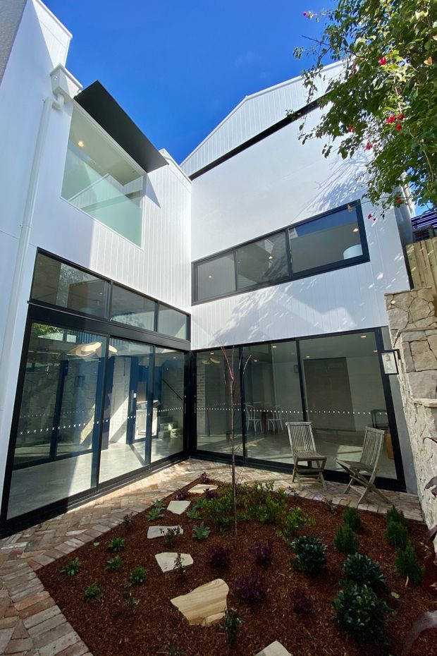 A bright, contemporary multi-level courtyard framed by white vertically ribbed wall cladding and extensive black-framed glazing. The enclosed courtyard contains brick paving, a landscaped garden bed with small shrubs, loose stepping stones, and simple outdoor timber chairs. A stone retaining wall and timber fence are visible at the side, with strong sunlight and clear blue sky above.