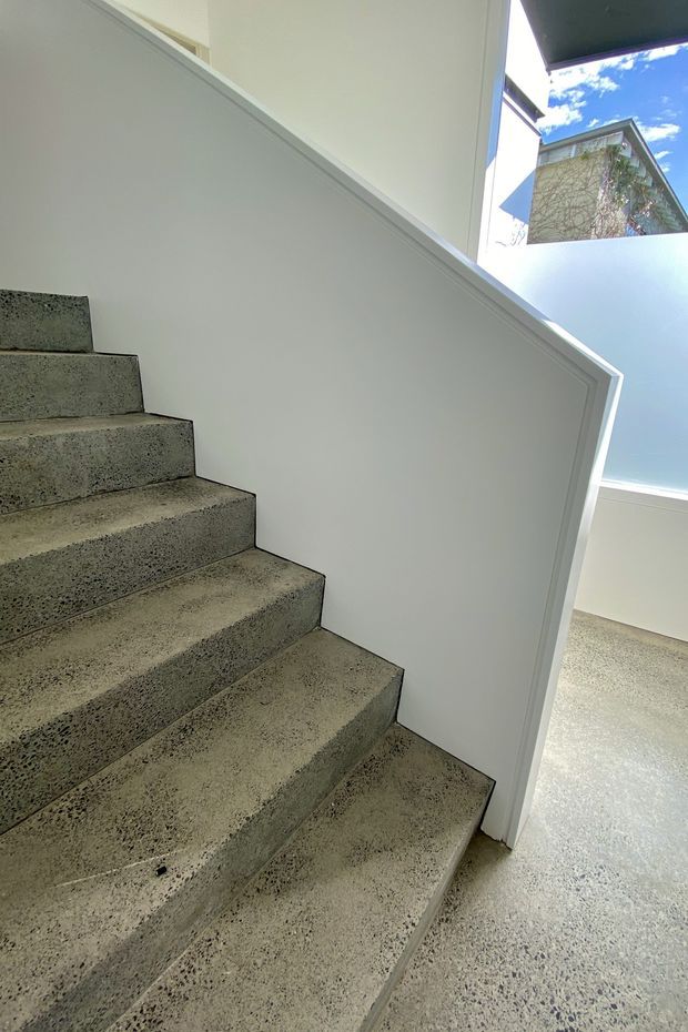 Interior view of a minimalist stairway with broad exposed aggregate concrete steps, a smooth white-painted stair wall and matching light-speckled concrete flooring. A bright glazed opening at the upper landing brings in daylight and offers a partial view of a neighbouring building and blue sky.