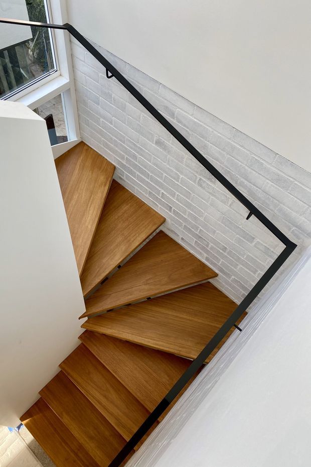 Interior view looking down a compact stairway with warm timber treads, a slim black metal handrail and balustrade, white-painted brick walls, and a tall window bringing in natural light. The stair geometry includes a turning or angular arrangement around the wall.