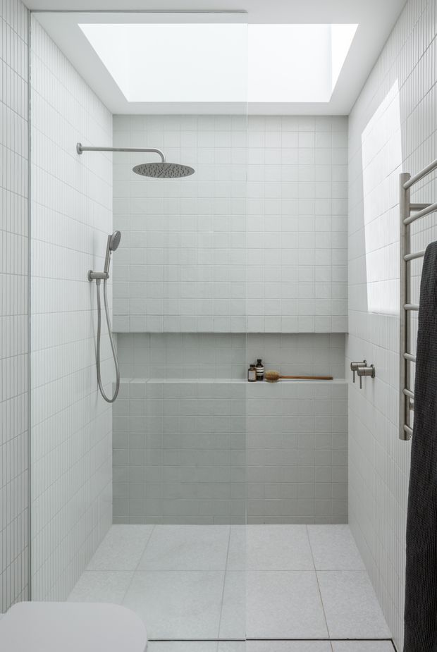 A bright, compact walk-in shower room with pale grey-white square tiled walls and floor, a large frameless skylight, rainfall and handheld shower fittings, a built-in tiled shelf, and a wall-mounted heated towel rail. The space has a clean, minimal, spa-like appearance with soft grey fixtures and limited warm timber accents.