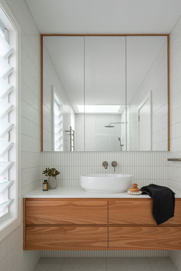 Bright contemporary bathroom vanity with a large frameless mirrored cabinet edged in timber. The room has white vertically lined wall tiles, a white integrated vessel-style basin, brushed metal wall-mounted tapware, warm timber floating drawers, a grey tiled floor, and a louvred window providing natural light. A shower with a skylight is visible in the mirror reflection.