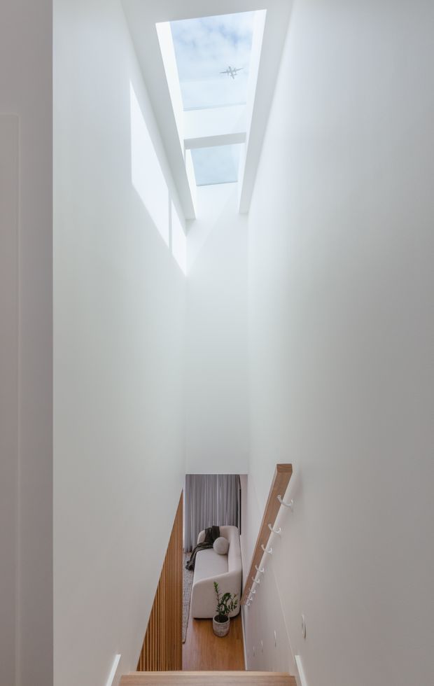 A narrow, tall stairway or landing with crisp white walls and a high ceiling fitted with multiple skylights. Strong daylight creates geometric shadows on the walls. A timber stair handrail with white hooks descends toward a lower-level living area, where a light upholstered sofa, timber flooring, curtains, and a small potted plant are visible.