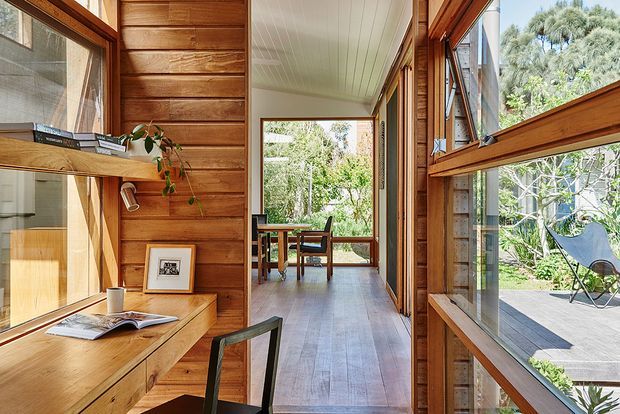 A warm timber-lined interior corridor or study nook with extensive glazing, built-in timber desk and shelving on the left, and a view through to a dining area and outdoor deck. The space features exposed timber wall panelling, timber-framed windows, a light painted plank ceiling, and timber flooring, creating a bright connection between interior and garden.