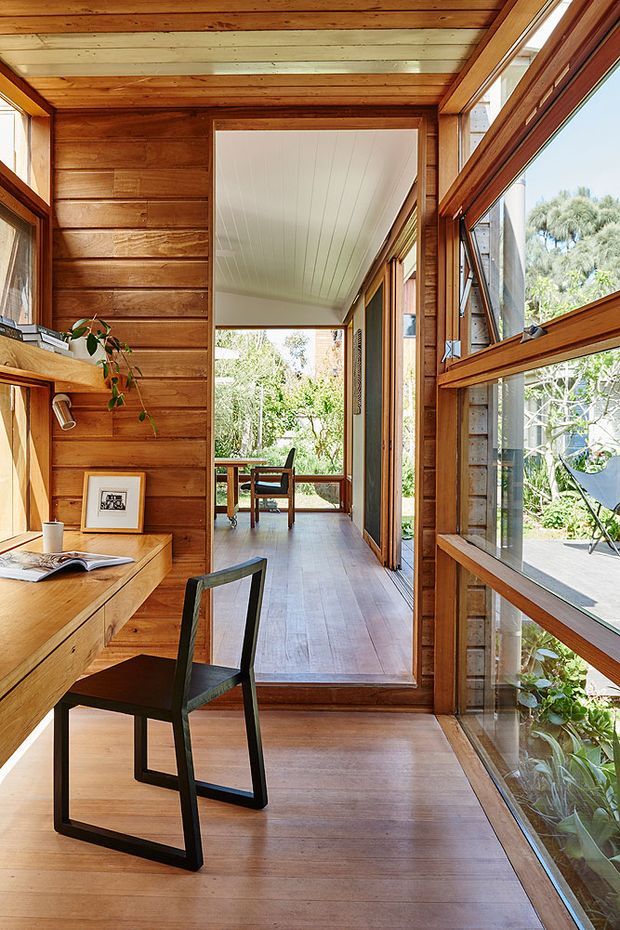 A warm timber-lined interior corridor and home office nook with extensive windows. The foreground contains a built-in timber desk with a dark-framed chair, while the corridor leads to a small sitting or dining area opening toward a leafy garden. Timber flooring, horizontal timber wall boards, timber window frames, and a white-lined ceiling create a light-filled, connected indoor-outdoor space.