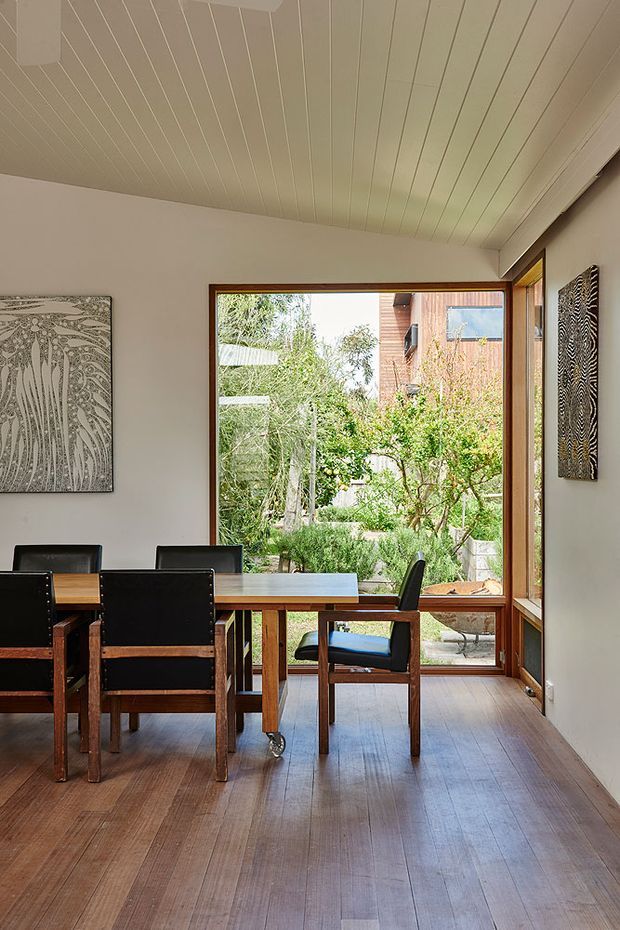 A warm, light-filled dining room with a timber dining table and black timber dining chairs, set beside large timber-framed windows overlooking a leafy garden. The room features wide-plank timber flooring, white walls, a white-lined ceiling with narrow boards, timber window frames, and monochrome patterned artwork.