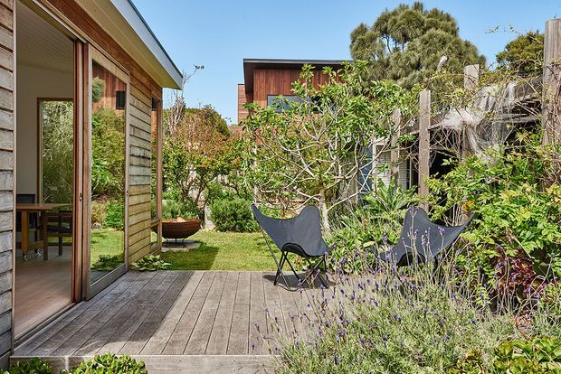 A sunny landscaped courtyard or deck between timber-clad structures. The foreground features a weathered timber deck, large glazed timber-framed sliding doors, and two black sling chairs surrounded by dense coastal-style planting. Additional timber-clad buildings and a sheltered outdoor area are visible beyond the garden.